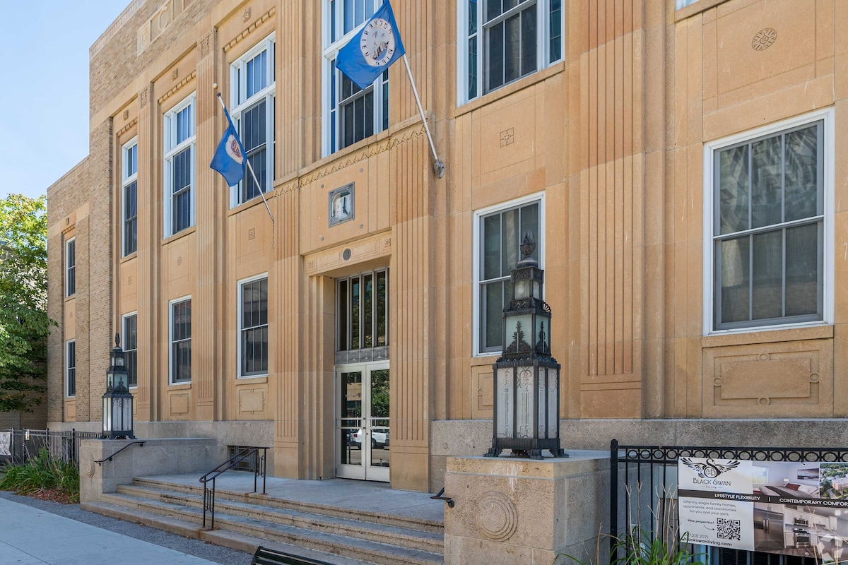 The historic exterior of the Old City Hall Apartments features classic architectural details, including prominently placed stone accents and large windows. Flags are displayed on either side of the entrance, and decorative lanterns flank the steps leading to the front door.