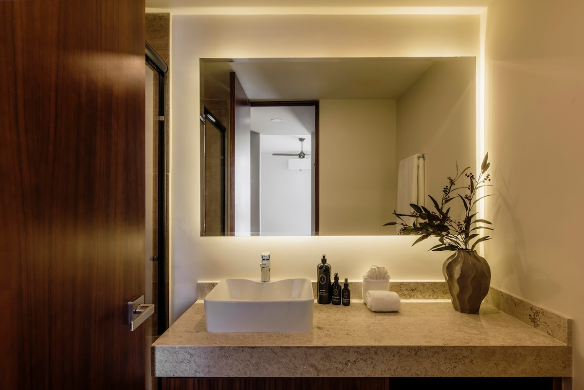 A modern bathroom features a sleek countertop with a spacious basin. A well-lit mirror is framed by warm ambient lighting. Decorative elements include a small vase with greenery and neatly arranged toiletries, providing a functional and stylish space.