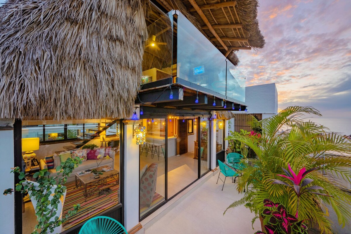 A two-level penthouse features a spacious terrace adorned with vibrant turquoise chairs and tropical plants. Floor-to-ceiling glass doors provide a view into the modern living area, where warm lighting illuminates the colorful decor. The palapa roof adds a unique tropical touch to the structure.
