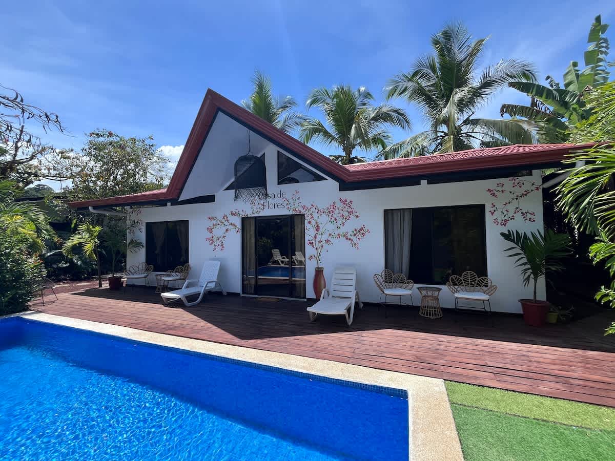 The exterior of the Casa de Flores is showcased, featuring a white facade adorned with artistic floral designs. A blue swimming pool is positioned in front, with white lounge chairs placed nearby. Lush greenery and palm trees surround the area, enhancing the serene setting.