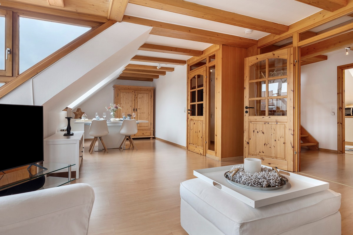 An open-plan living space is showcased with wooden beams above, enhancing the natural light that fills the area. A cozy sofa is positioned in front of a television, while a dining table with two chairs is visible in the background. Light-colored wooden furniture adds a warm touch.