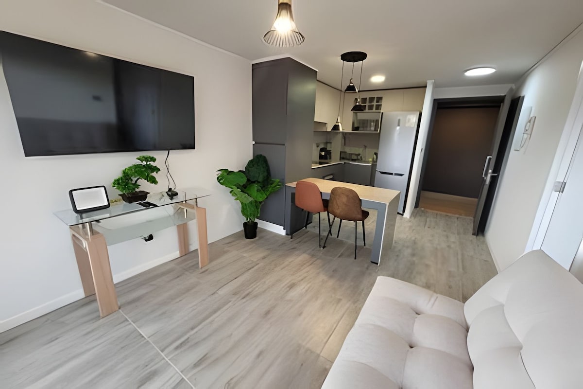 A modern living space features a large flat-screen TV mounted on the wall, while a cozy beige sofa sits nearby. A dining table is surrounded by two chairs, and compact kitchen appliances are visible in the background. Decorative plants add a touch of greenery.