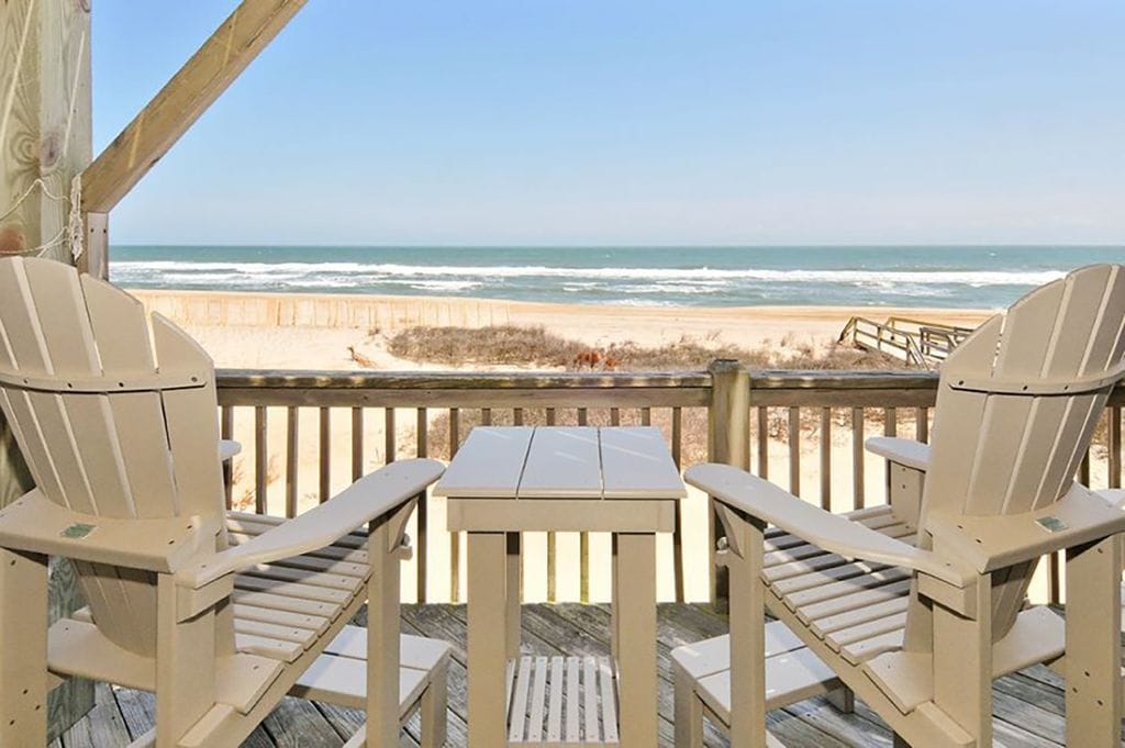 A beachfront deck is shown with two beige Adirondack chairs positioned around a small table. The ocean view features gentle waves and a sandy shore, with a picturesque wooden railing framing the scene.