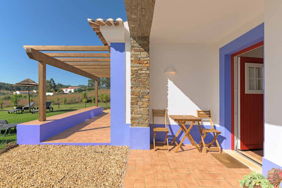 An inviting outdoor seating area features a small wooden table and two folding chairs under a pergola. The space is bordered by a gravel pathway and stretches towards a green lawn, enhancing accessibility to the surrounding garden area.