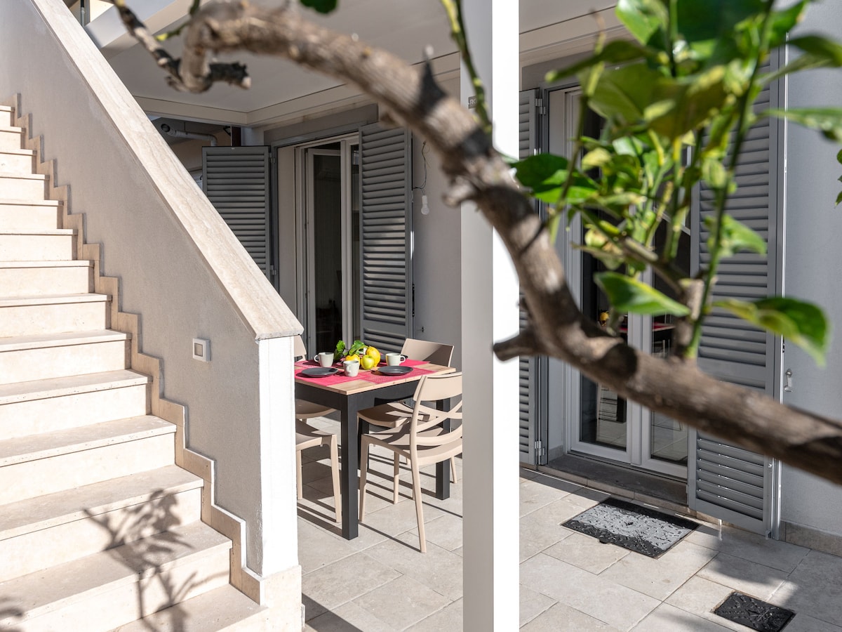 A dining area is visible under a covered terrace, featuring a table set with bright dishes and chairs. Stairs ascend to another level, while sliding glass doors with shutters provide access to the indoor space. A leafy branch frames the scene.