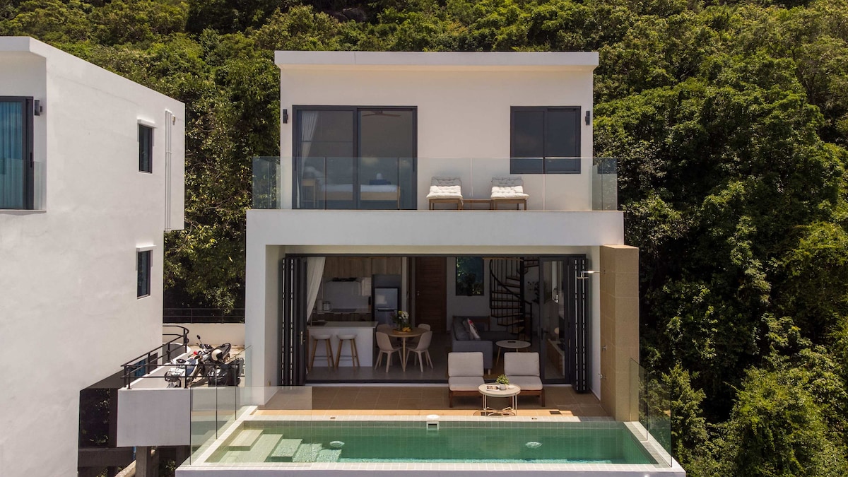A modern villa is showcased from an aerial perspective, characterized by large glass sliding doors that open to a private pool area. The upper terrace features two sunbeds, while lush greenery forms a backdrop, contributing to the tranquility of the setting.