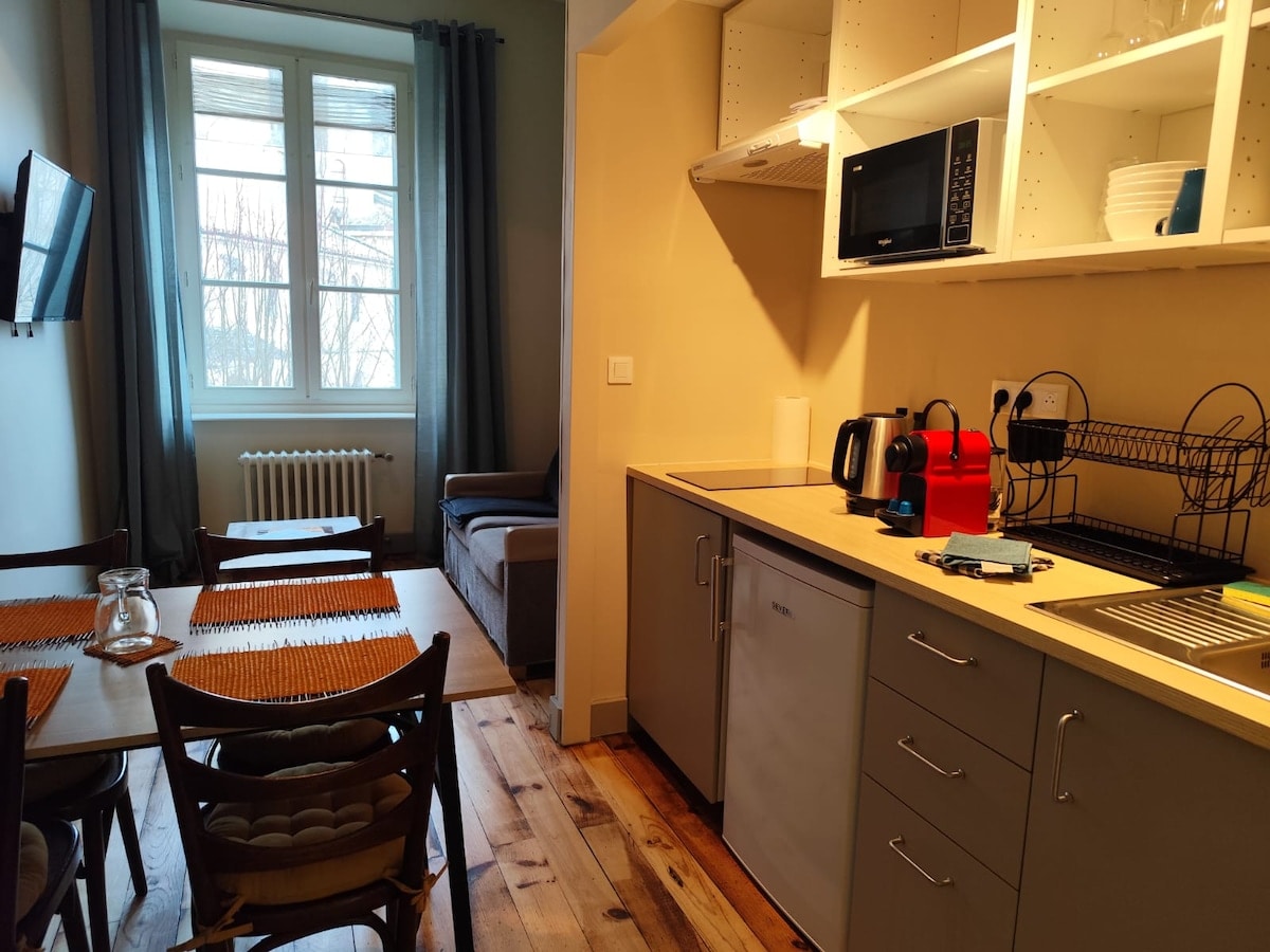 A modern kitchen area is displayed with gray cabinets and a countertop. A microwave, kettle, and refrigerator are visible, along with a dark dish rack. A dining table with chairs is present, and a sofa is partially seen in the background near a window framed by light curtains.