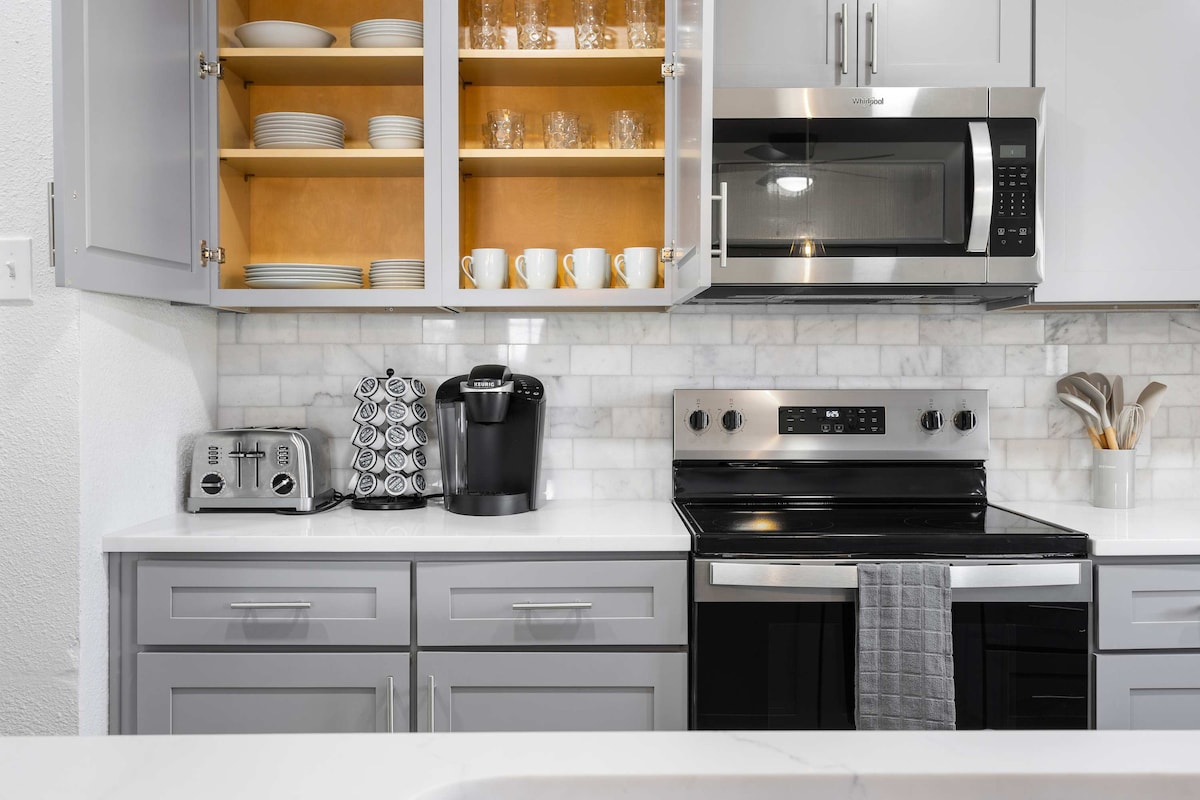 The kitchen features modern appliances, including a stainless-steel microwave and oven. Shelves hold neatly arranged dishes and glassware, while a coffee maker is positioned beside a stylish toaster. Light-colored cabinets provide a clean and bright appearance against the marble backsplash.