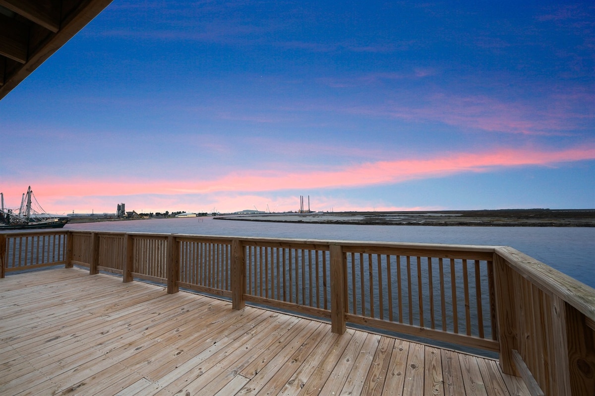 A wooden deck offers views of the calm water at sunset, with soft pink and blue hues gracing the sky. The surrounding landscape includes distant structures and wetlands, emphasizing the tranquil setting.