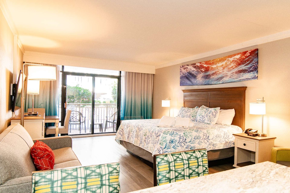 A spacious studio room includes a cozy king-sized bed with floral bedding, a comfortable gray sofa, and a kitchenette with seating at the breakfast bar. Large glass doors provide access to a private balcony, with fresh natural light enhancing the vibrant decor.