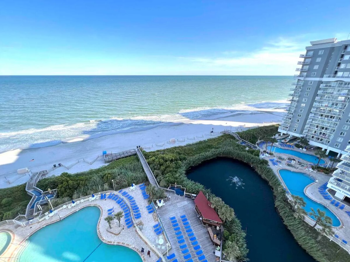 An expansive view of the ocean showcases gentle waves and a sandy shore. The image includes resort swimming pools and lush greenery surrounding a lagoon, with sun loungers placed invitingly around the pool area, creating a tranquil beachside environment.