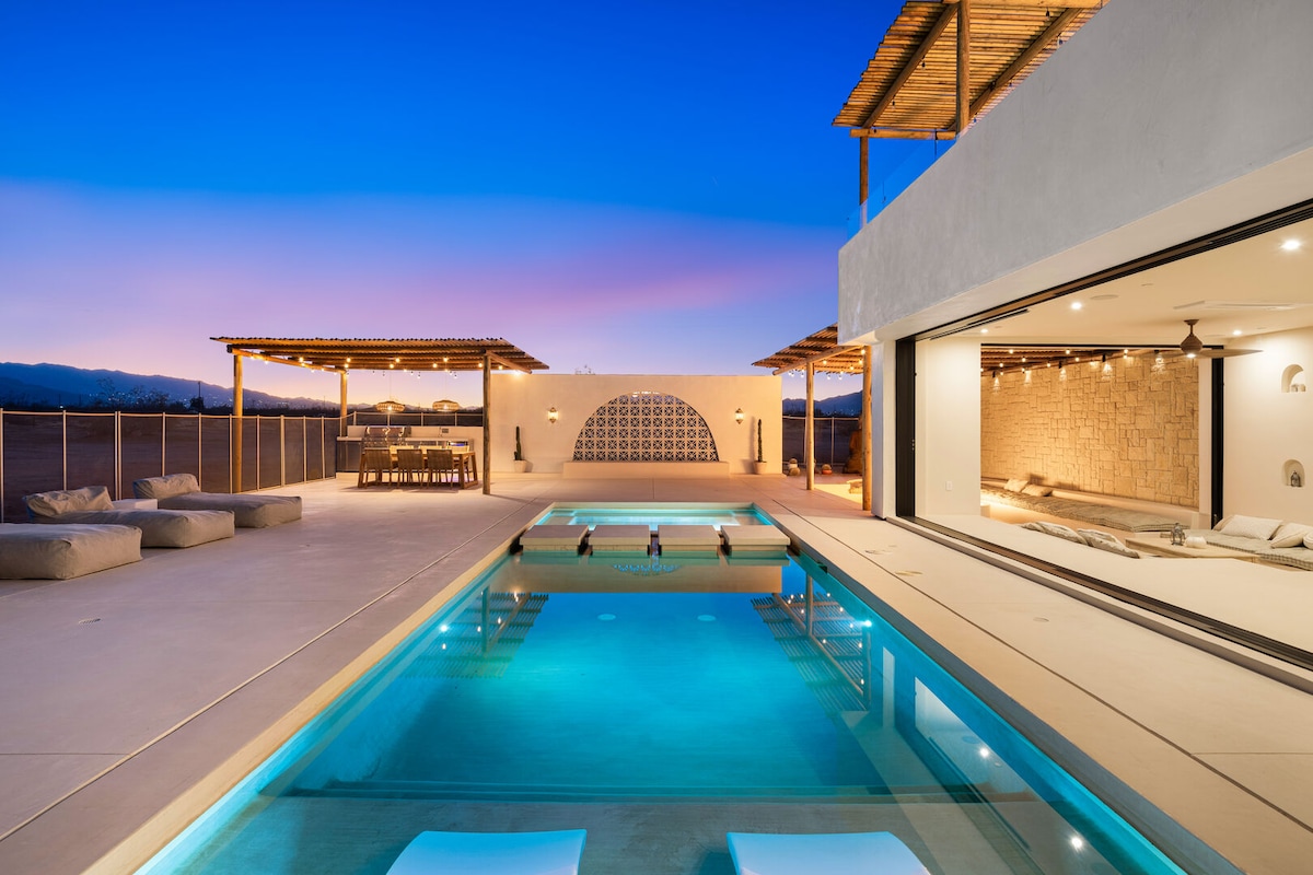 A tranquil resort-style pool reflects the evening sky, surrounded by a spacious patio that offers loungers and a shaded dining area. The open-concept living space features large glass doors, allowing for a seamless transition between indoors and outdoors.