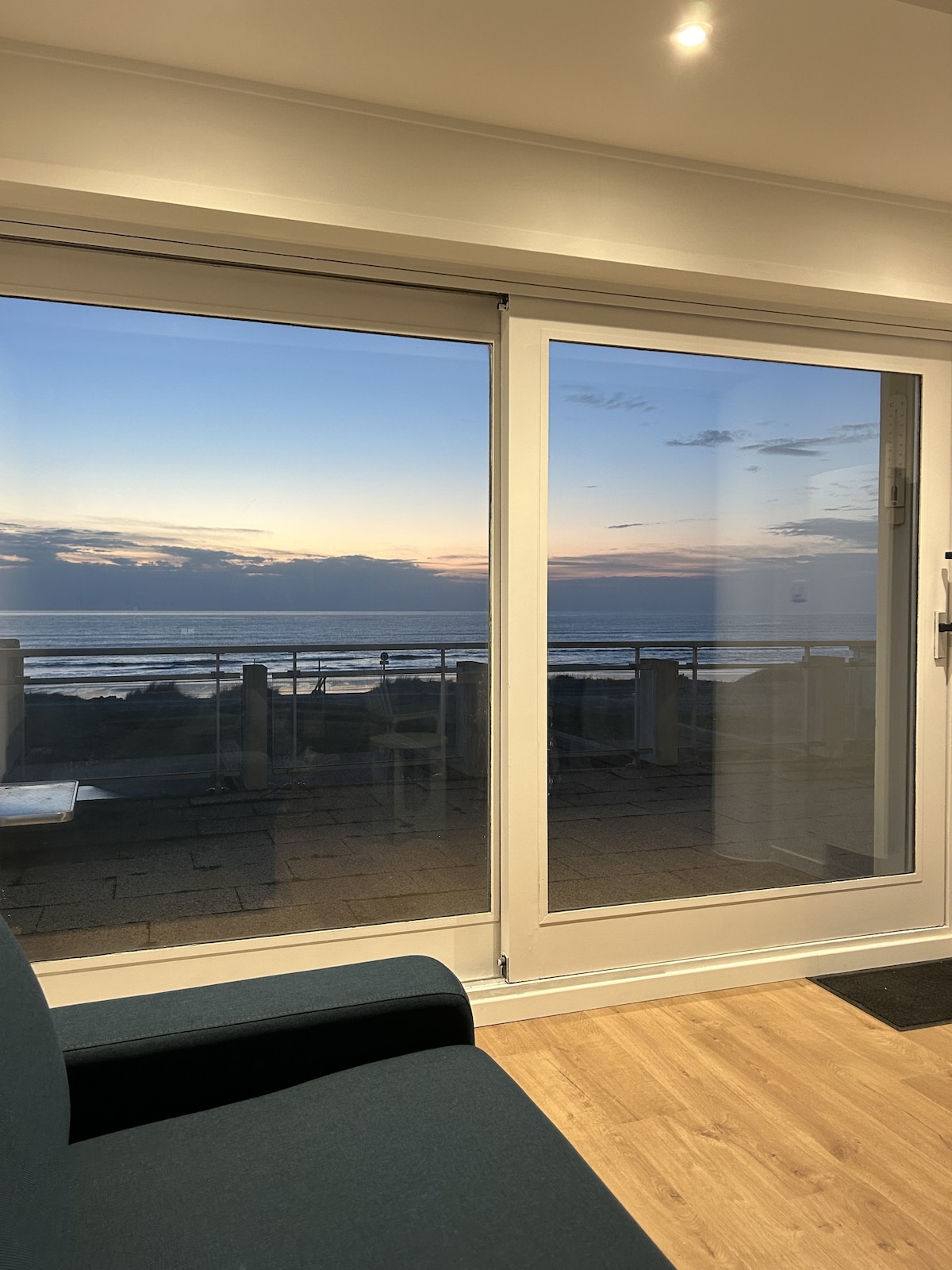 Floor-to-ceiling windows provide an unobstructed view of the sea at sunset. The calming colors of the sky reflect on the water, enhancing the serene ambiance of the living space, while a cozy sofa is positioned nearby.