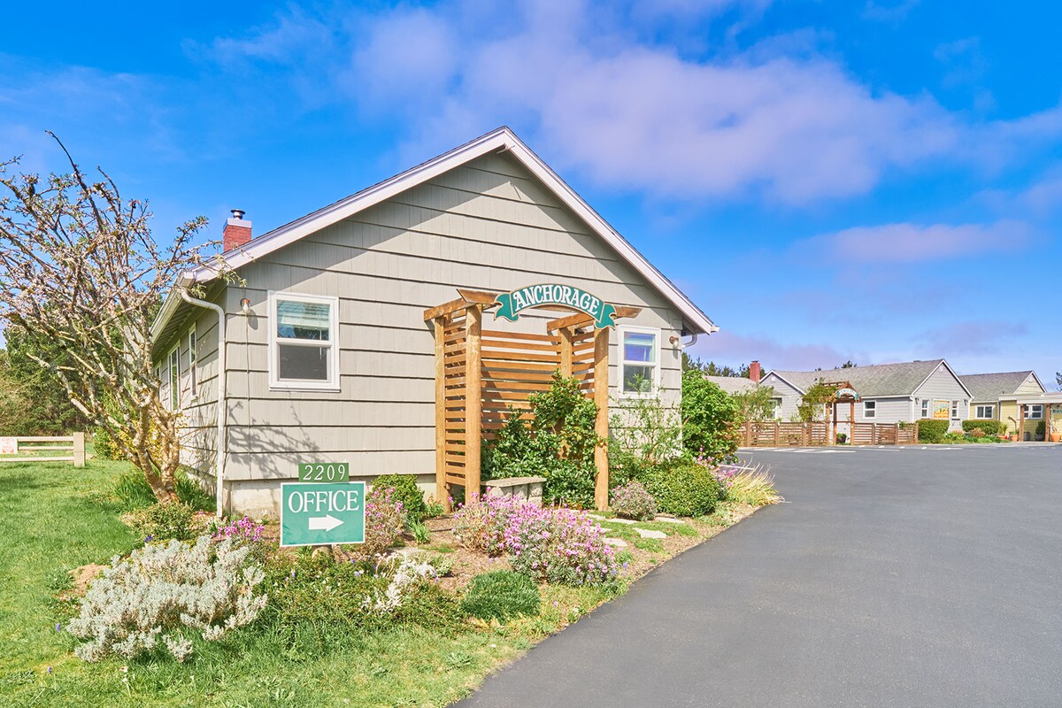 The exterior of Anchorage Cottages #4 is portrayed, featuring a welcoming entrance with a wooden arbor. Surrounding vibrant gardens display a variety of flowers and greenery. A sign indicating the office is visible in front of the building, which has a light grey facade and a charming roof.