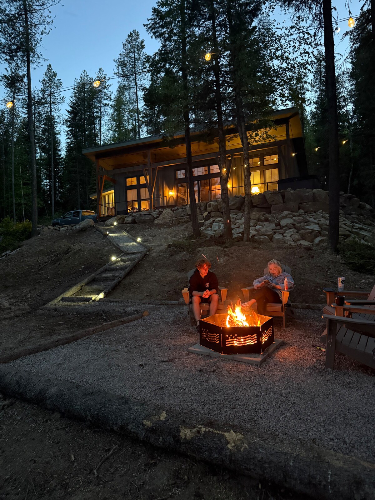 A warm glow emanates from the house, surrounded by tall trees and starlit skies. Two individuals sit by a firepit, nestled on outdoor chairs. The pathway leading to the house is illuminated, highlighting the rock steps that connect the fire area to the entrance.