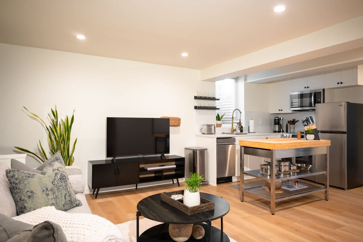 A modern living space features a comfortable sofa and a sleek TV stand, complemented by a circular coffee table. The kitchenette boasts stainless steel appliances and a wooden island, while natural light filters through a nearby window, enhancing the contemporary design.