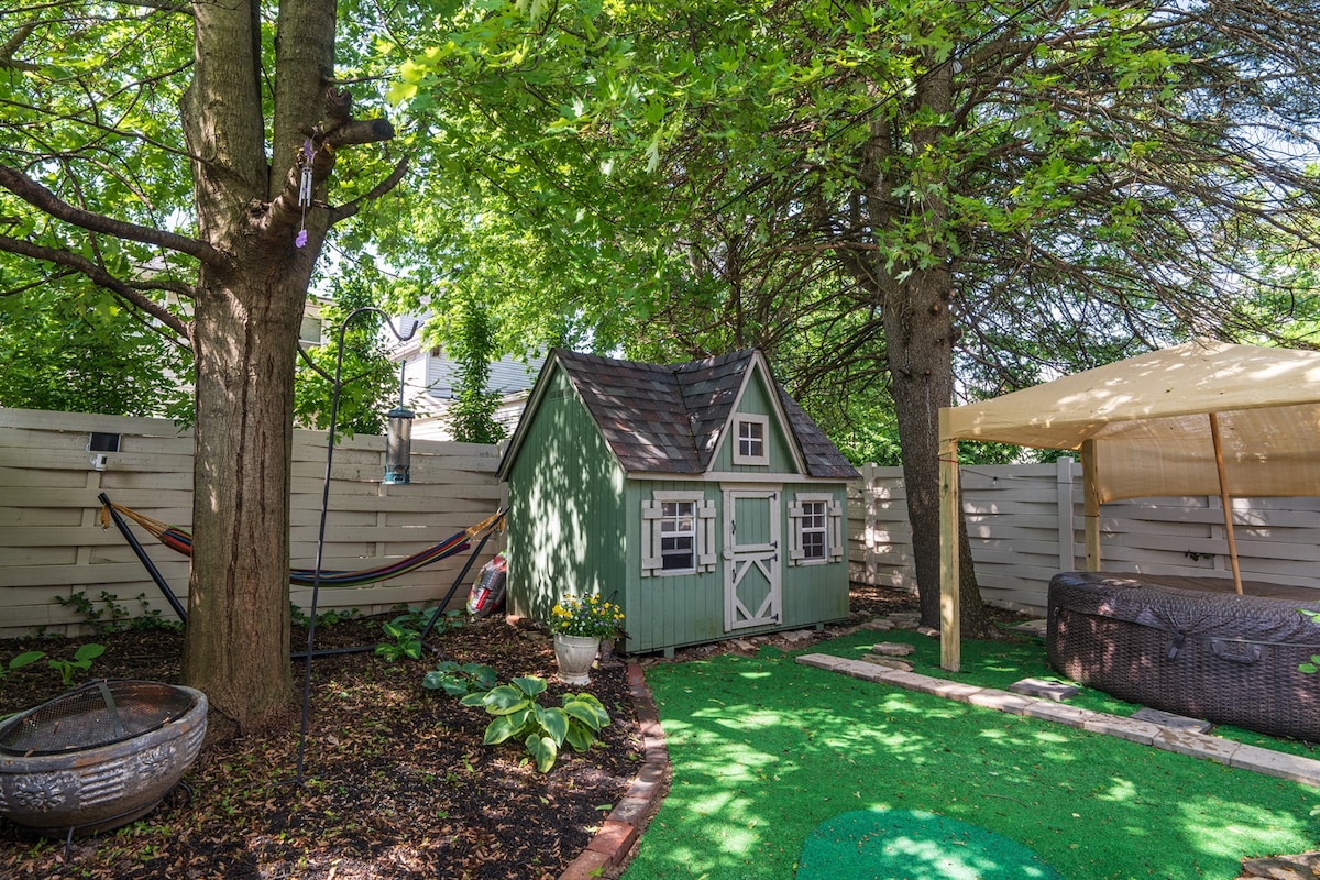 The fenced backyard features a children's play shed styled in green, surrounded by lush greenery and decorative stones. A hammock is visible, along with a covered seating area. The soft turf creates an inviting atmosphere, complemented by vibrant trees and landscaping.