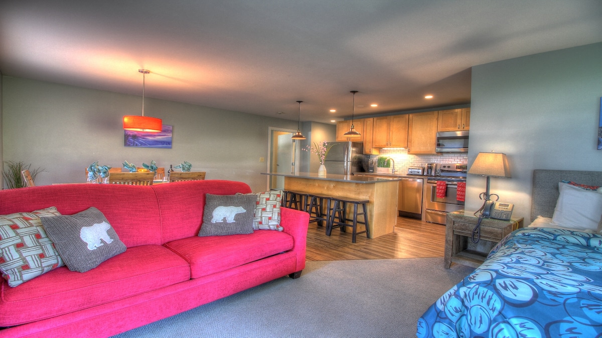 An open living space features a bright red sofa with decorative pillows, complemented by a warm wood dining area with stools. The kitchen, equipped with stainless steel appliances, is visible in the background. Soft lighting creates a welcoming ambiance throughout the suite.