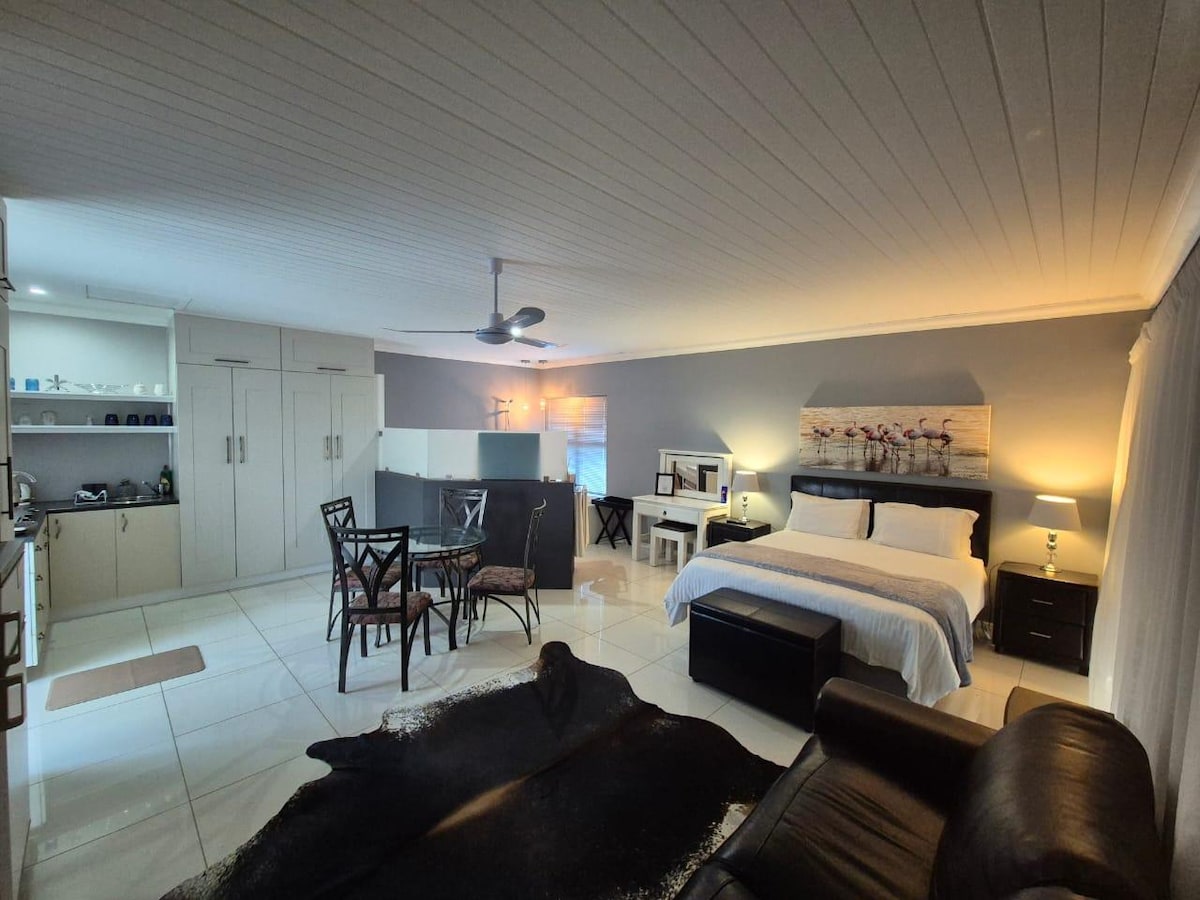 An open-plan living space contains a spacious bed with white linens and a dark bench at its foot. A kitchenette is visible in the background, with a dining area featuring a table and chairs. Ambient lighting creates a calm atmosphere.