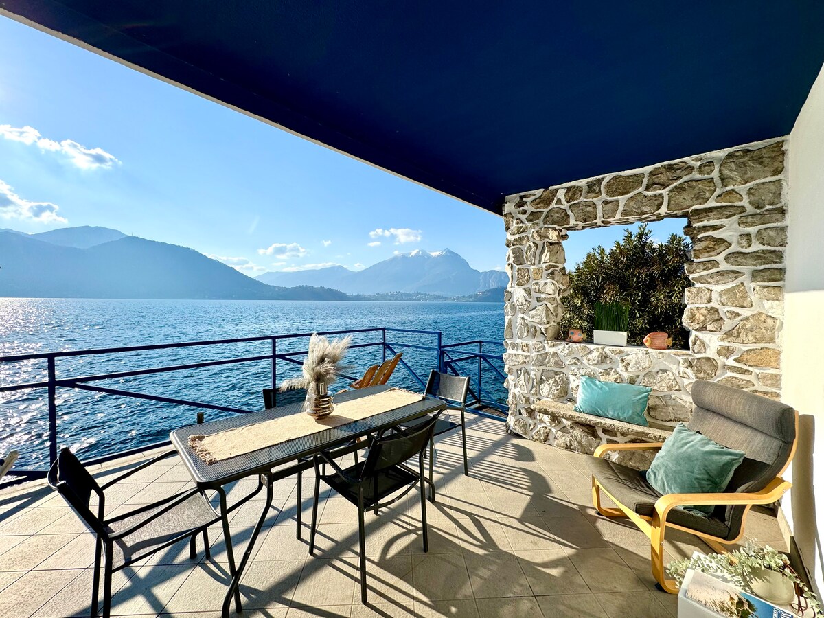 An outdoor terrace features a dining table with comfortable chairs, overlooking the shimmering waters of Lake Como. The surrounding mountains create a serene backdrop, while decorative items add a touch of style. Soft cushions invite relaxation, enhanced by the bright sunlight filtering through.
