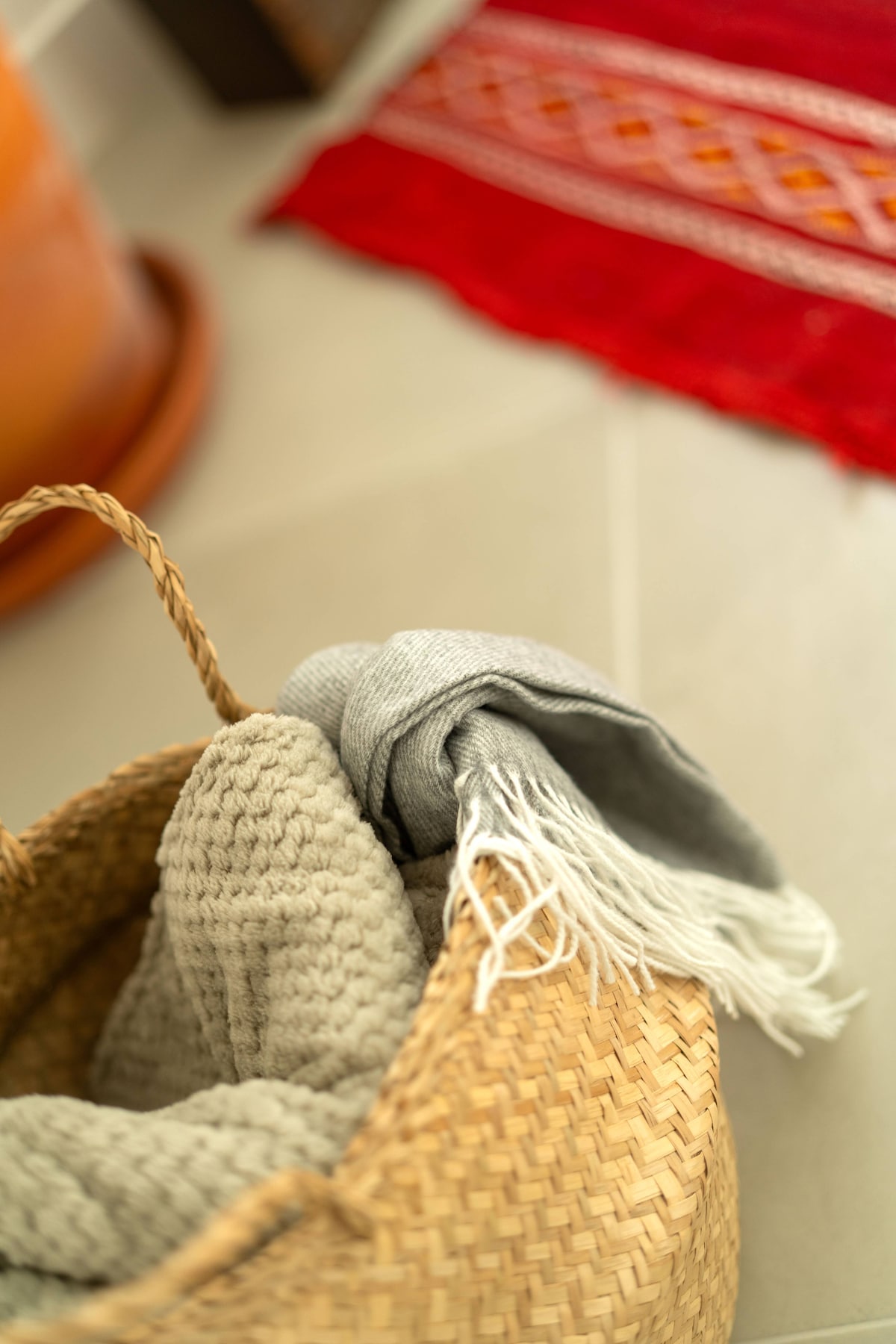 A woven basket contains soft, textured grey blankets, with fringed edges partially draped over the rim. The basket sits on a tiled floor, near a vibrant red rug featuring intricate patterns.