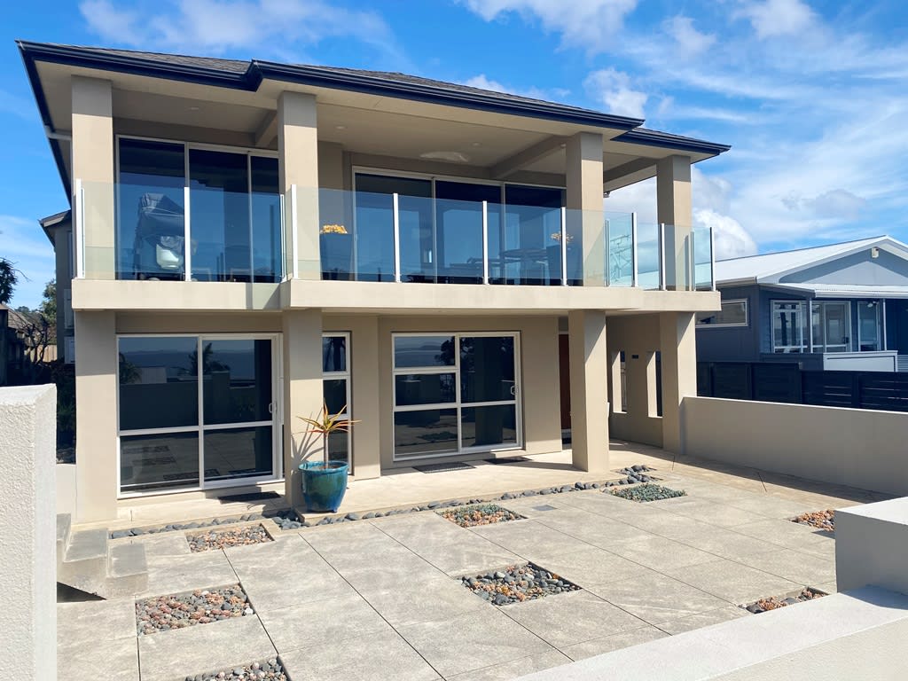 The modern two-story home features a spacious front terrace paved with large stones, accompanied by decorative gardens. Expansive glass panels on the upper floor offer unobstructed views, while the entry is framed by sleek architectural lines under a clear blue sky.