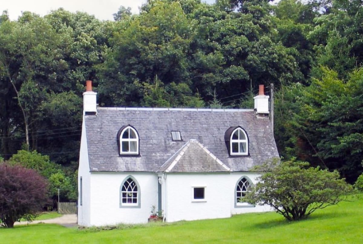 A charming cottage with a steeply pitched roof and two prominent gable windows is surrounded by lush greenery. The white exterior is softened by the natural landscape and features a manicured grassy area in the foreground, enhancing the peaceful setting.
