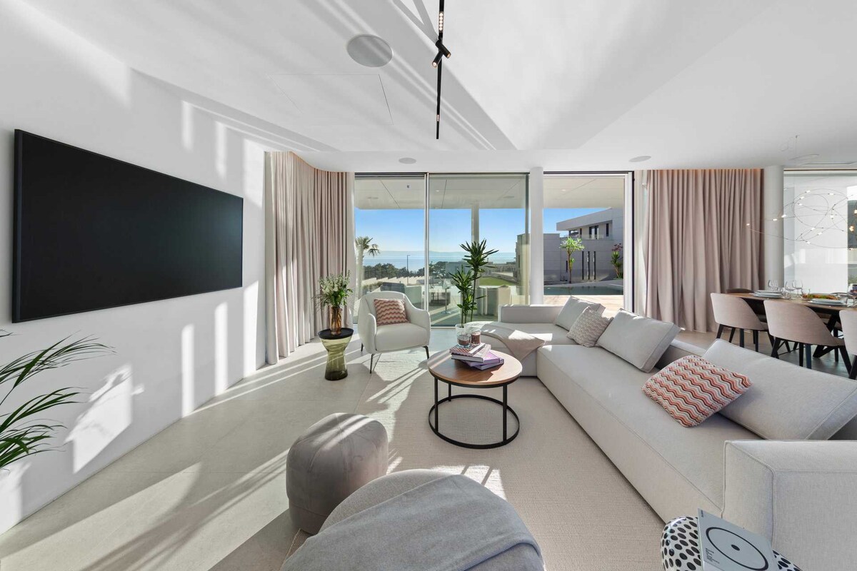 A bright living area features sliding glass doors that offer outdoor access and views of the surrounding greenery. A modern sectional sofa is complemented by accent pillows and a circular coffee table. Natural light filters through light drapes, creating a welcoming atmosphere.