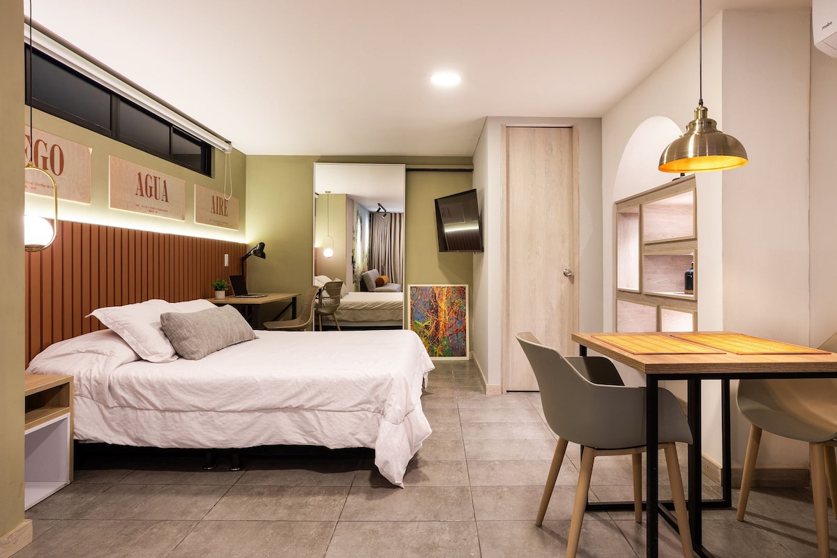 A well-lit loft room features a spacious bed against a textured wall. A dining area with a wooden table and modern chairs is visible, along with a seating area that includes a mounted television. A hallway leading to amenities, including a closet, can be seen in the background.