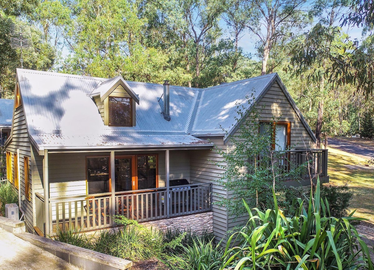 The exterior of the Blackwood Chalet is surrounded by trees, showcasing a multi-level structure with a sloped roof. Large windows are seen along the front, providing natural light, while a spacious deck area features railing and a barbecue. Lush greenery complements the home's aesthetic.