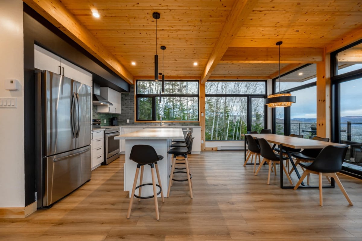 A modern kitchen and dining area are highlighted by large windows that showcase views of the surrounding nature. A central island with seating complements high-quality appliances. Wooden beams and light-colored walls create an airy atmosphere, while a spacious dining table awaits gatherings.