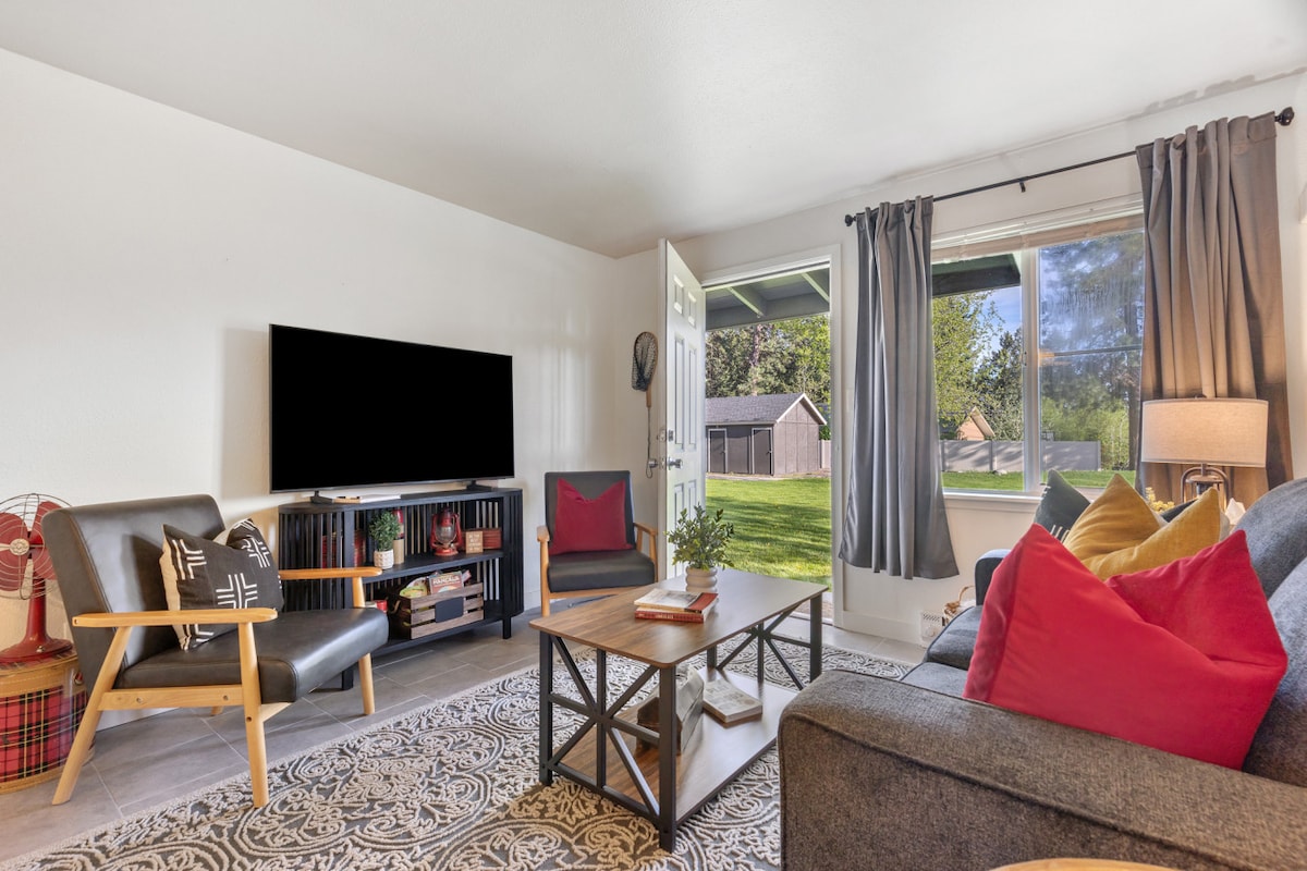 A modern living area features a large smart TV mounted on a white wall, complemented by a gray sofa adorned with red and black accent pillows. A wooden coffee table sits in the center, surrounded by two comfortable chairs. Natural light streams in from a nearby window, enhancing the welcoming space.