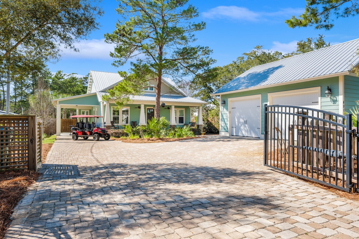 A charming beach home is seen with a modern exterior, featuring a blue façade and metal roof under a clear sky. Paved stones create a spacious driveway leading to a garage and parking area, with a golf cart parked alongside. Lush greenery surrounds the property.