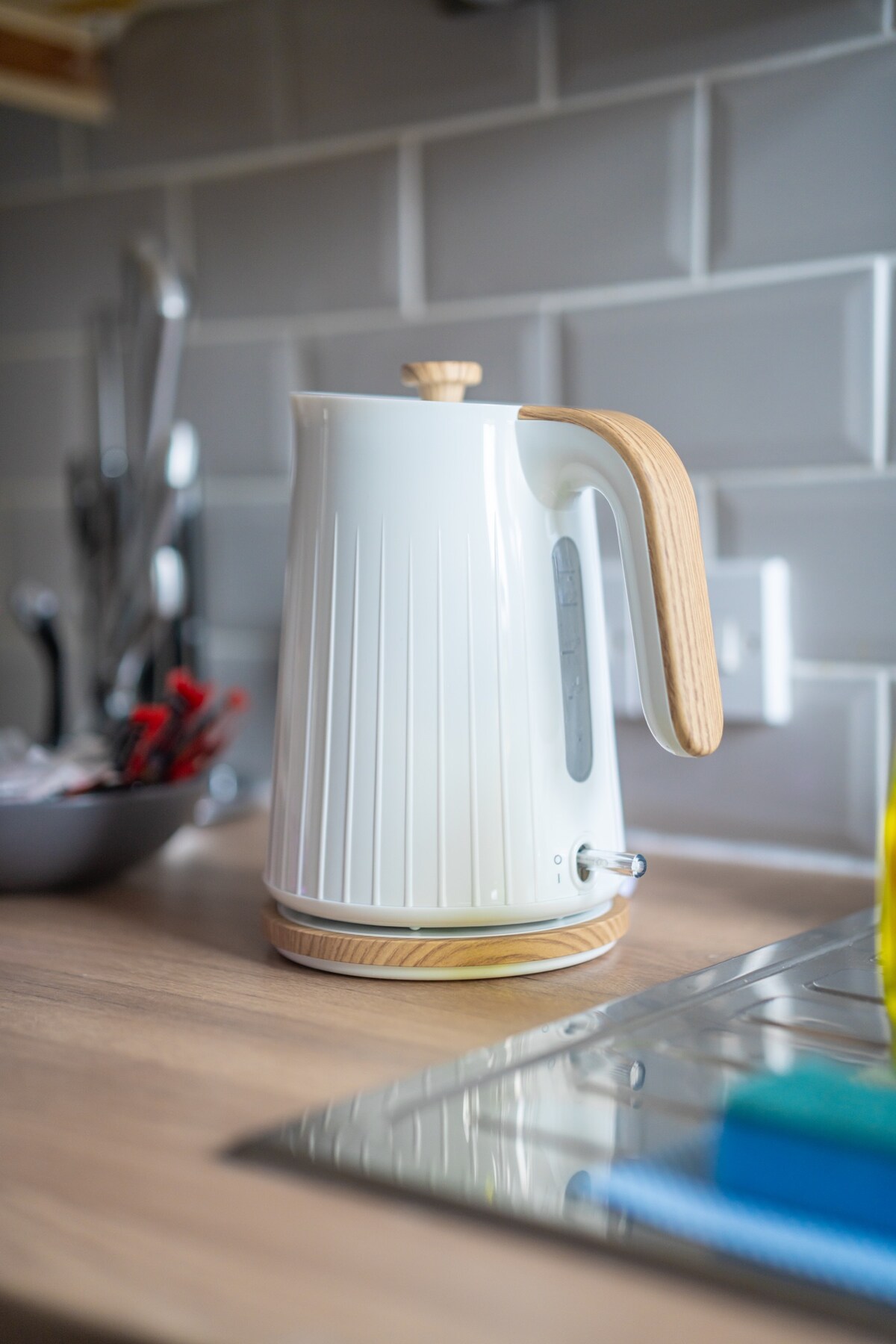 A modern electric kettle is positioned on a wooden countertop, featuring a sleek white body and a natural wooden handle. The kettle's transparent water level indicator is visible, along with a light grey tiled backsplash that complements the kitchen's contemporary design.