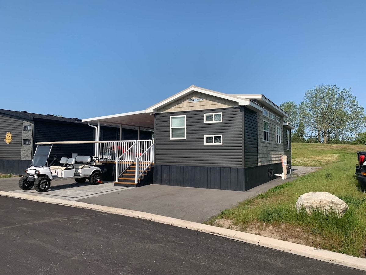 The exterior of a modern cottage is showcased, featuring dark siding and a welcoming entrance with steps leading to the door. A golf cart is parked beside the unit, under a covered area, while green grass and trees are visible in the background.