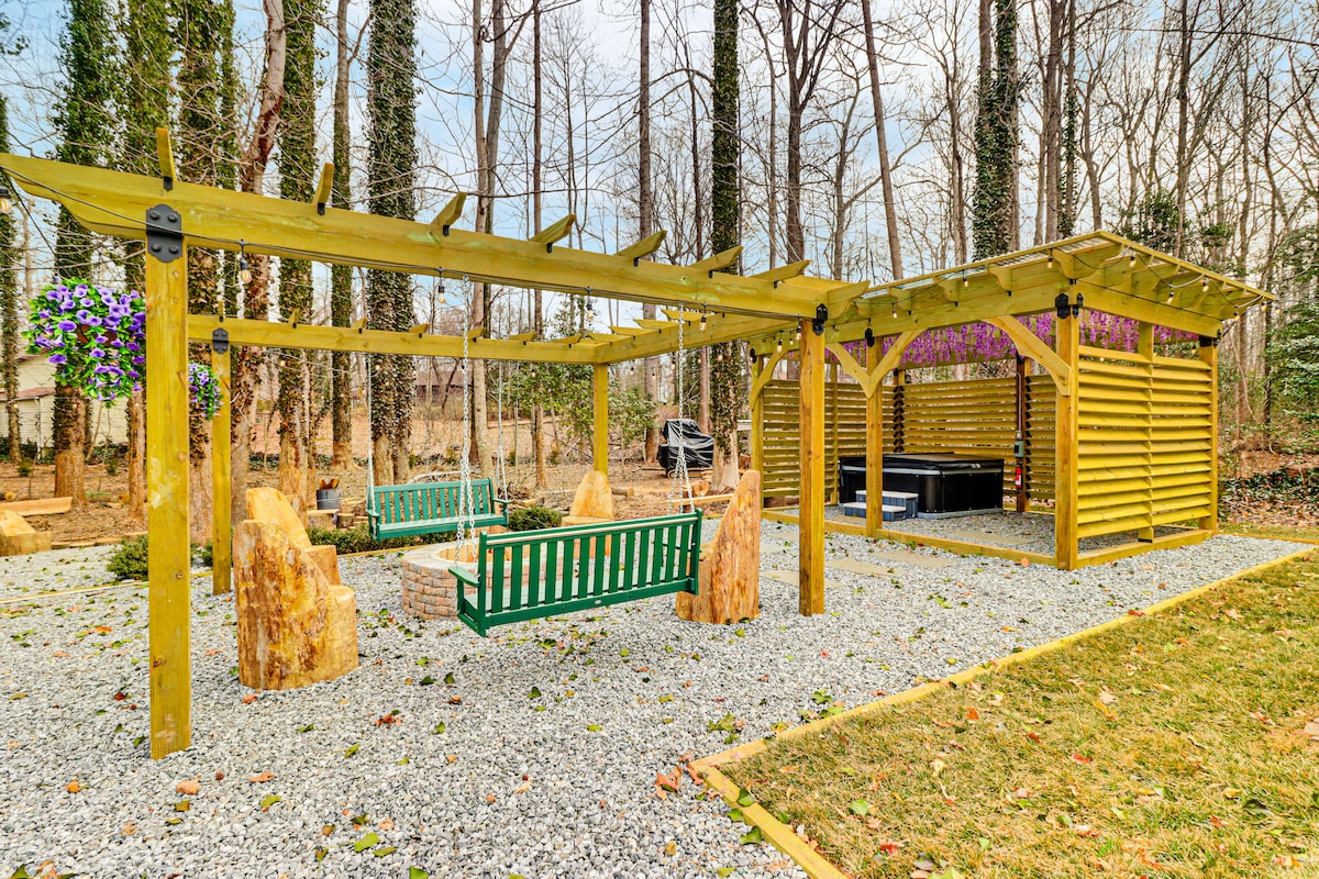 An outdoor area features a wooden pergola with swings and benches crafted from natural materials. Gravel and grass encompass the space, with a hot tub partially visible under the pergola, surrounded by trees that provide a sense of privacy.