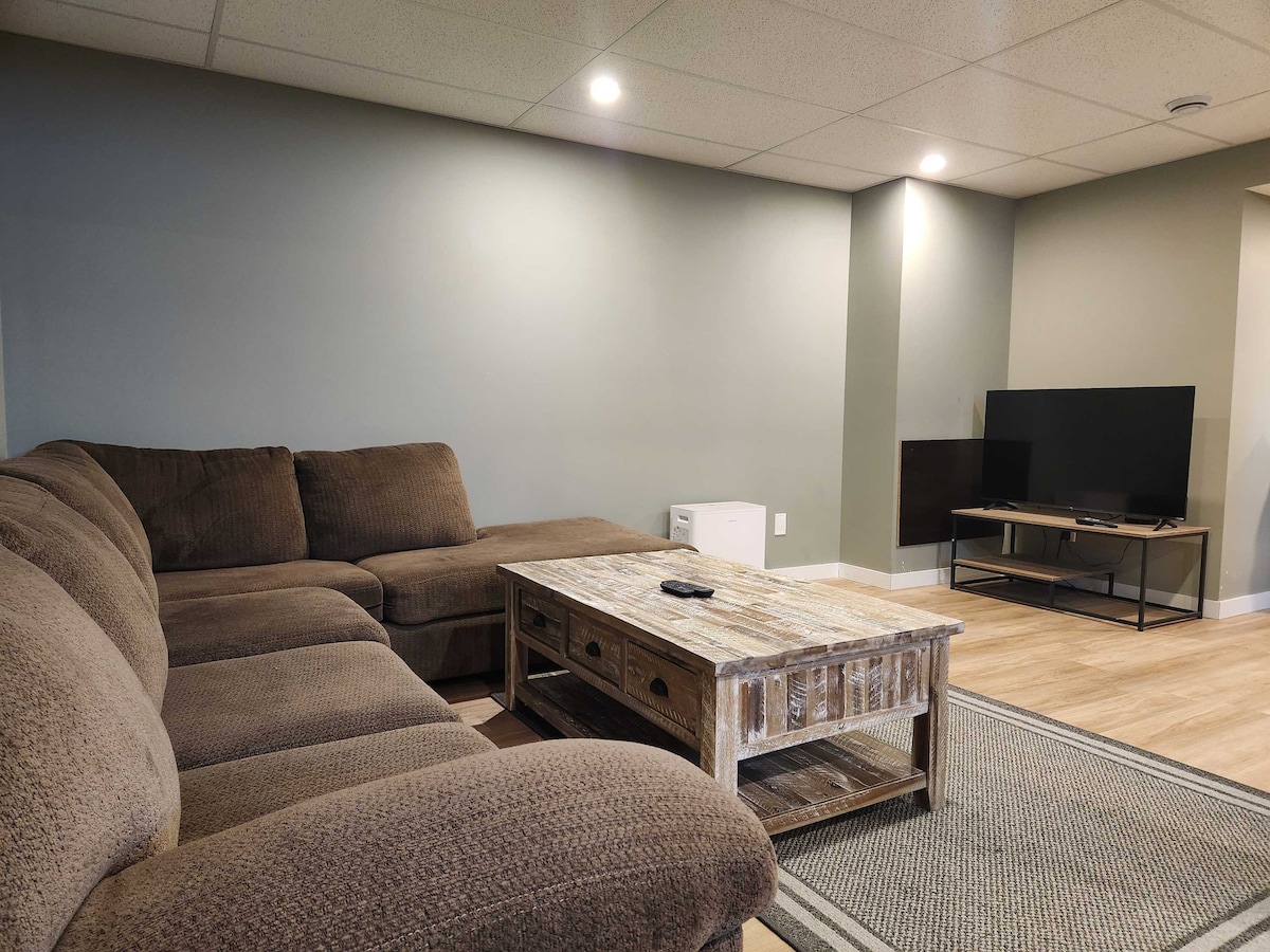A spacious living area features a large sectional couch upholstered in a soft brown fabric, paired with a rustic coffee table. A flat-screen TV is positioned on a low stand, and warm lighting enhances the homey feel, complemented by light wood flooring.