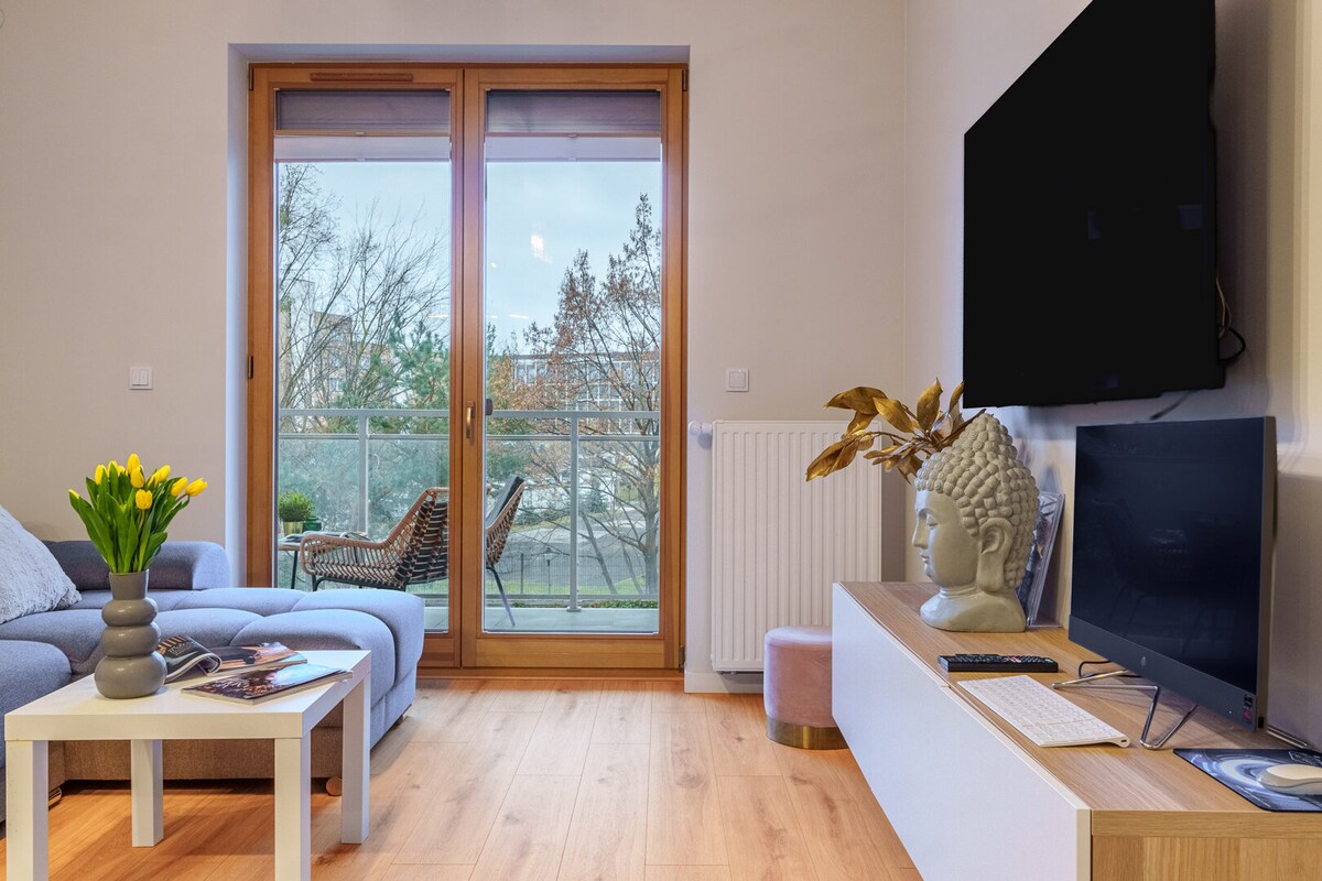 A light-filled living area features a soft blue sofa, a coffee table, and a flat-screen television. Large glass doors open to a balcony, revealing tranquil outdoor seating and leafy views. A vase with fresh yellow tulips adds a touch of color to the space.
