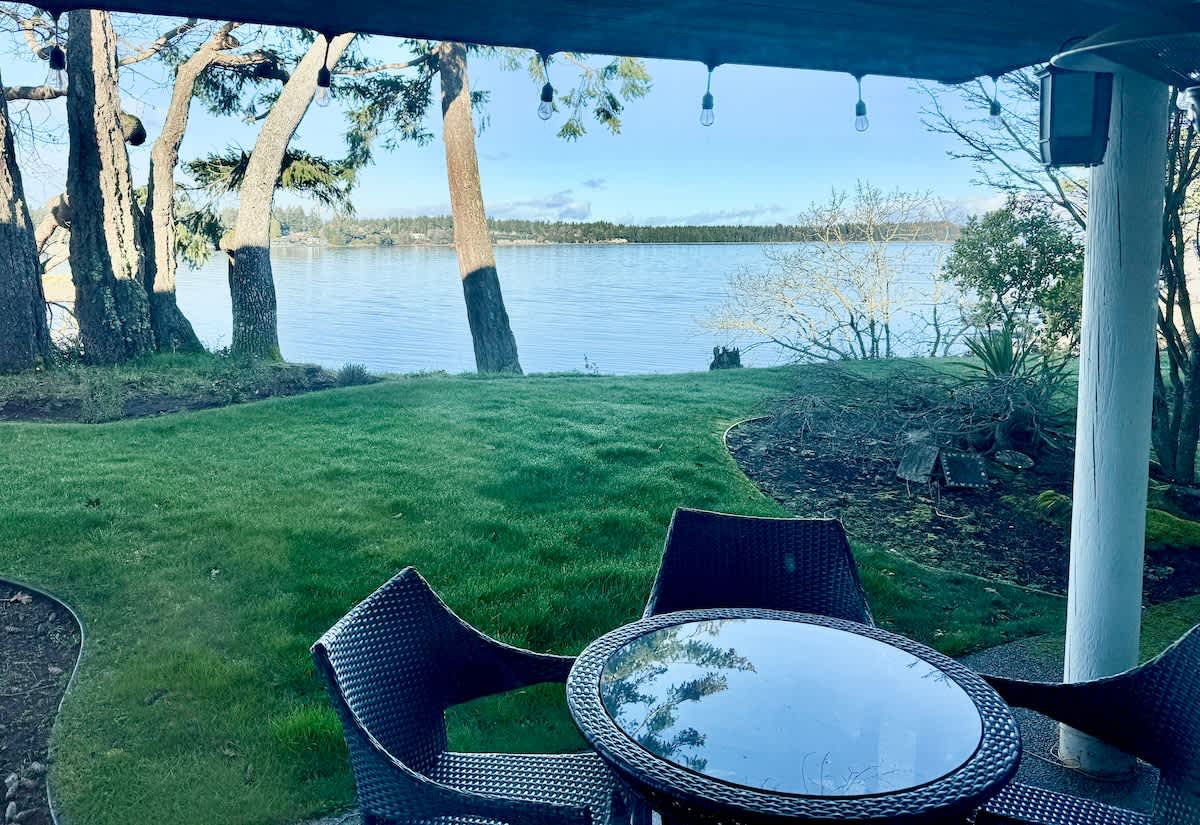 A tranquil view is captured from a covered patio, featuring a round glass table surrounded by four dark wicker chairs. Lush green grass extends toward a calm waterway, framed by trees and distant shoreline under a clear sky.