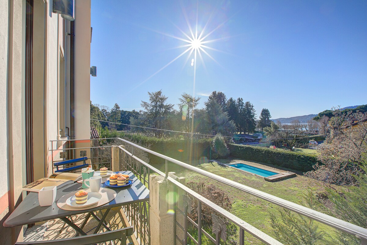 A balcony is equipped with a small table and chairs, presenting an inviting setting for al fresco dining. A spread of pastries and drinks is visible on the table. Sunlight bathes the area, and a swimming pool is seen in the garden below, surrounded by greenery.