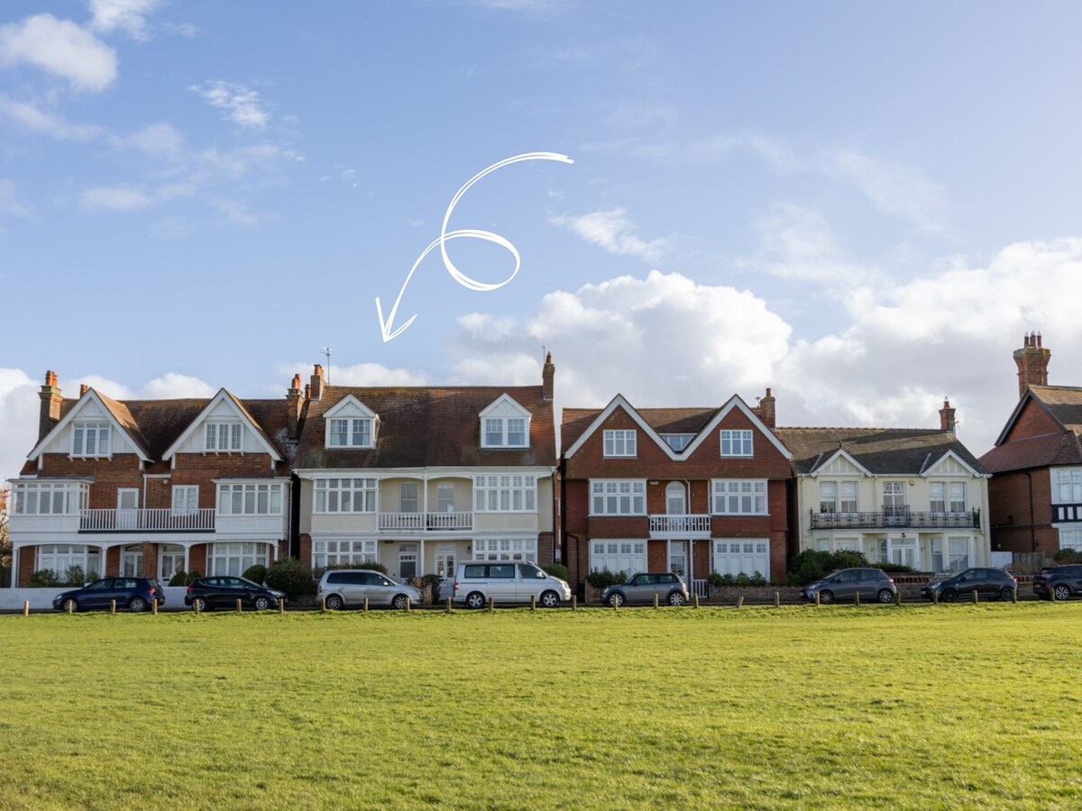 A row of traditional townhouses is visible, showcasing a charming architectural style with pitched roofs and gabled ends. The greenery of Southwold Common is situated in the foreground, with parked cars lining the street. A clear blue sky with scattered clouds enhances the overall scene.