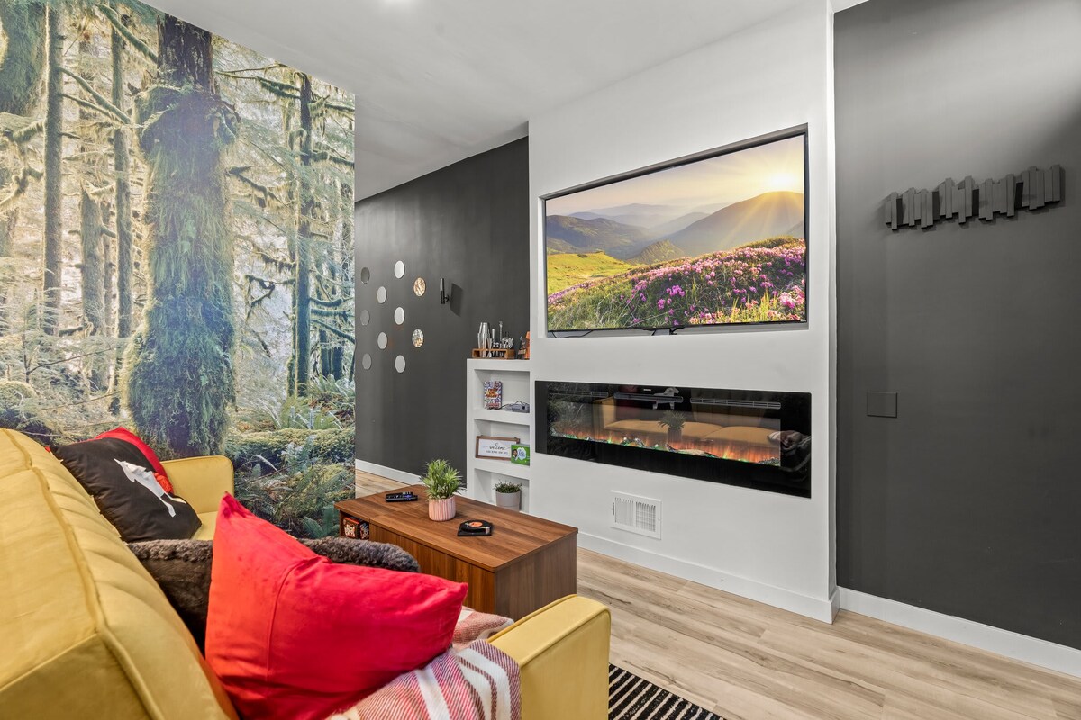 A comfortable yellow sofa adorned with decorative pillows and throws is set against stunning forest-themed wallpaper. A wall-mounted smart TV is positioned above a modern electric fireplace, enhancing the relaxing atmosphere of the living room.