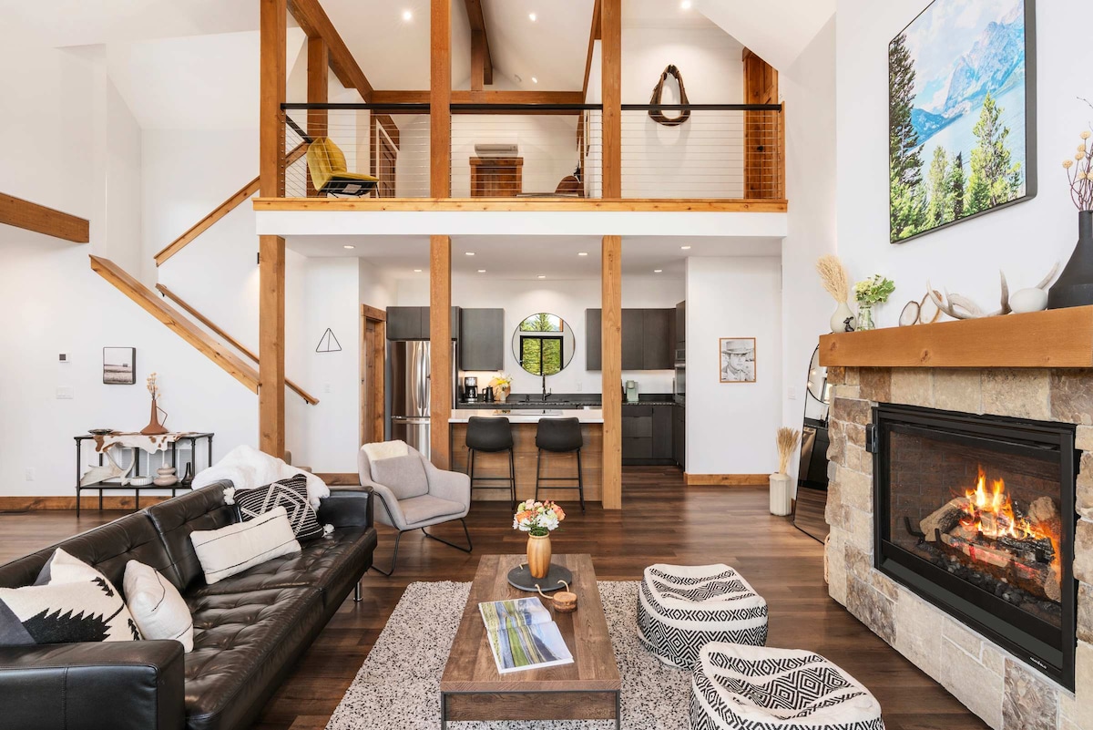 The living room features a comfortable leather sofa and a cozy gas fireplace. A wooden coffee table is surrounded by patterned poufs. The open design connects to the dining area, showcasing the modern kitchen and a staircase leading to the loft above.
