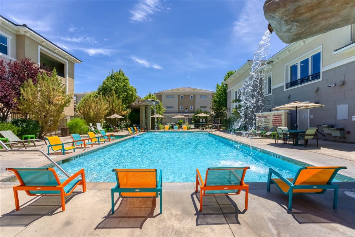 A spacious outdoor pool is surrounded by colorful lounge chairs in orange and teal. Umbrellas provide shade for guests at nearby seating areas. Lush greenery and decorative landscaping enhance the tranquil atmosphere, complemented by a water feature adding soothing sounds to the space.