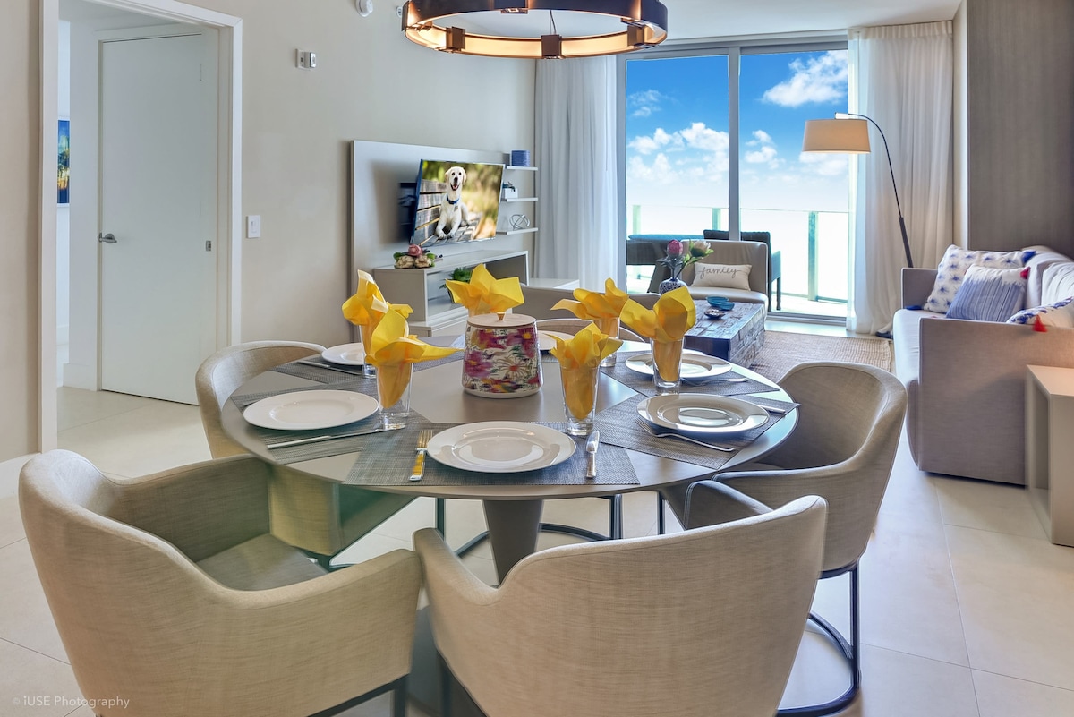 A round dining table is set for four, featuring yellow napkins and glasses filled with yellow flowers. A cozy living area is visible in the background, with a beige sofa and large floor-to-ceiling glass doors that offer ocean views. Natural light brightens the space.