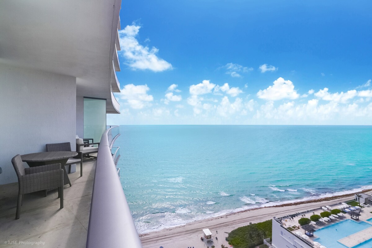 An ocean view is featured from a spacious balcony, showcasing clear blue waters and a bright sky. Outdoor furniture is visible, inviting relaxation while enjoying the scenery. The coastline is seen below, with beach amenities and lounge chairs positioned along the shore.