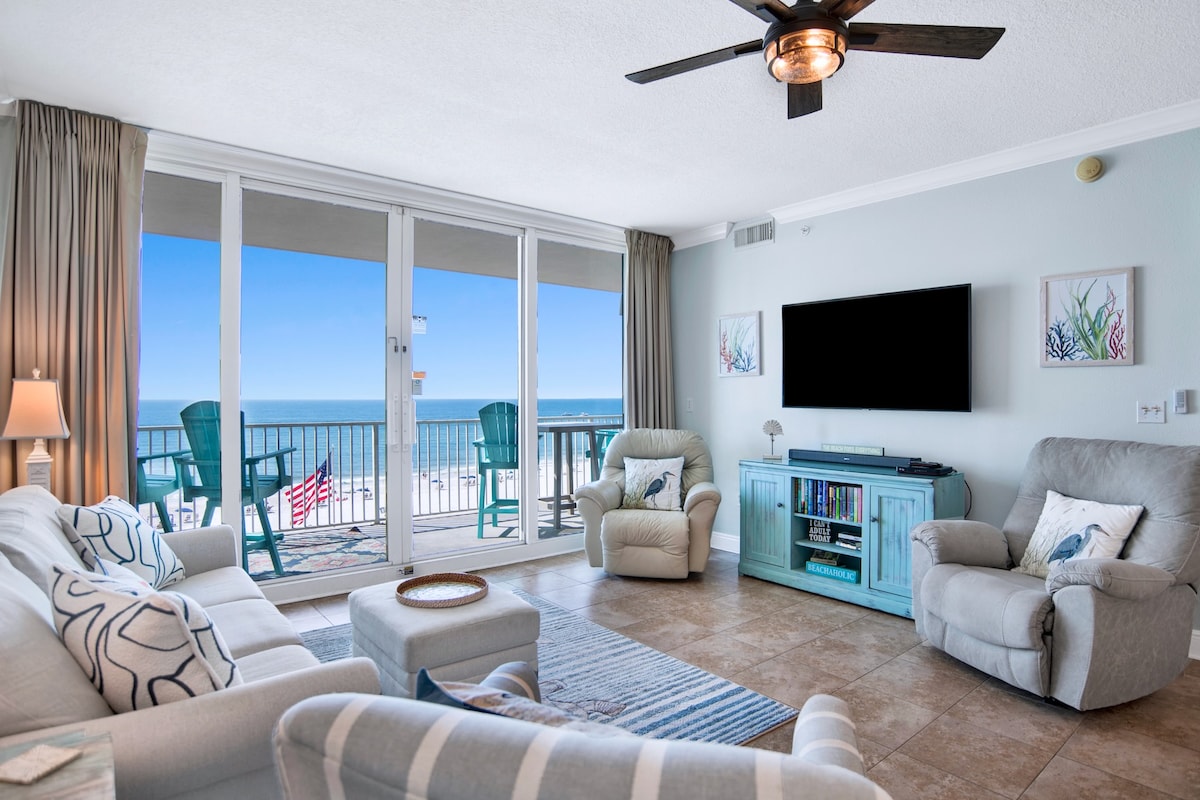 A bright living area is furnished with a light-colored sofa and recliners, complemented by coastal decor. Large glass doors provide direct views of the beach and ocean, while a flat-screen TV is mounted on the wall opposite the seating. Soft natural light fills the space.