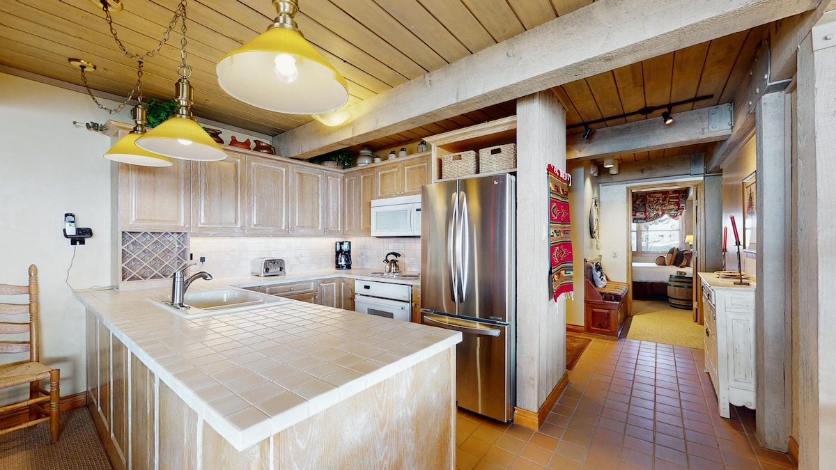 A spacious kitchen is equipped with modern appliances, including a stainless-steel refrigerator and a microwave. Cabinets are adorned with natural wood finishes, complemented by tile countertops. Bright overhead lights illuminate the space, while a view extends to an adjoining living area featuring a cozy seating arrangement.