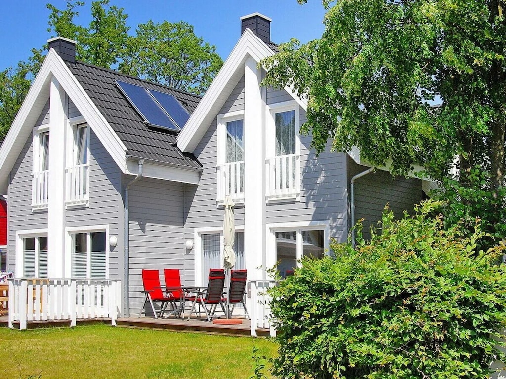 A semi-detached holiday home is presented in Scandinavian style. The exterior features a pale grey facade with white accents and balconies. A deck area with red outdoor chairs is visible, set against a backdrop of manicured greenery, creating a bright and inviting space.