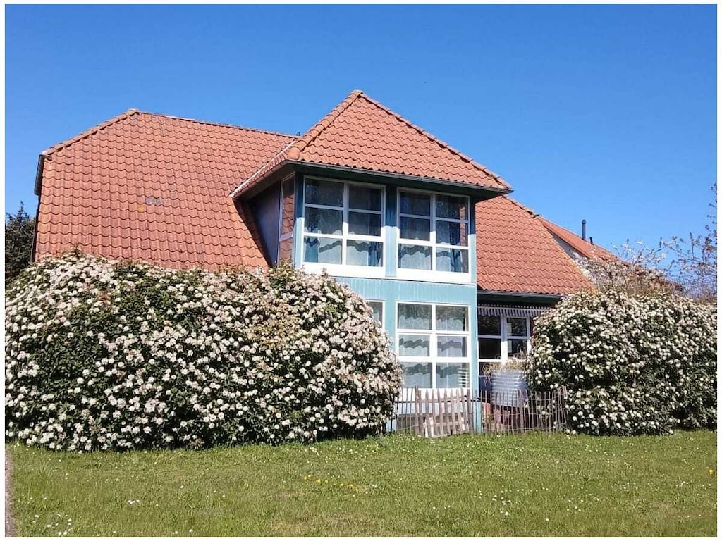 A charming two-story building features a sloped red tile roof and large windows adorned with light curtains. The exterior is surrounded by lush, flowering bushes, creating a vibrant contrast against the blue sky. A white picket fence adds a welcoming touch.